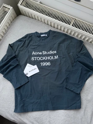 Mörkgrå långärmad Acne Studios tee - Snygg mörkgrå långärmad t-shirt från Acne Studios med vit text 'Acne Studios STOCKHOLM 1996' på bröstet. Tillverkad i mjuk bomull och har en relaxed passform. Perfekt för dig som gillar minimalistisk och stilren design.