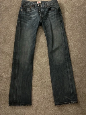 Levi's 501 raka jeans mörkblå - Klassiska Levi's 501 jeans i mörkblå denim med raka ben och knappgylf. Snygga kontrastsömmar och femficksmodell. Perfekta för dig som gillar tidlös stil och vill ha ett par slitstarka jeans i bomull.