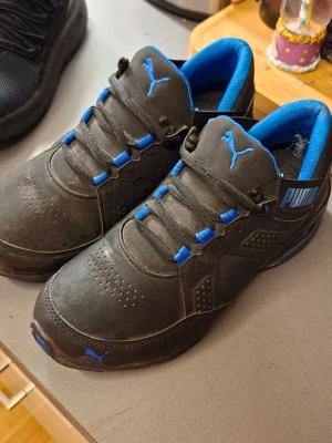 Svarta Puma sneakers med blå detaljer - Snygga svarta sneakers från Puma med blå detaljer på plösen, snörningen och insidan. Skorna har en låg profil och är tillverkade i syntetmaterial med perforerade partier framtill för extra ventilation. Klassisk Puma-logga på sidan och tån.