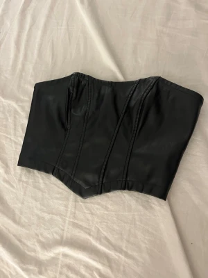 Svart läderlook bustierkorsett - Snygg svart korsett i läderimitation med markerade sömmar och dragkedja bak. Axelbandslös design med lätt formande effekt och stilren look. Perfekt för att skapa en edgy outfit eller som statement-plagg.
