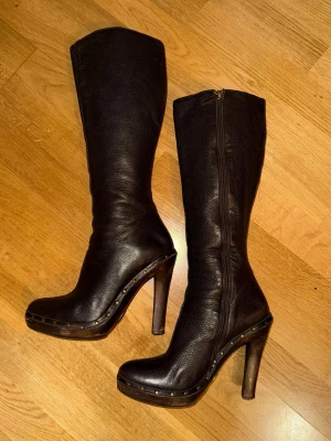 Vintage Gucci Mörkbruna höga läderstövlar med trä klack - Säljer ett par vintage Gucci mörkbruna höga stövlar i äkta läder med rundad tå och trä platform klack. Snygga nitar längs sulkanten och dragkedja på insidan. 