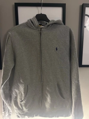 Grå hoodie från Polo Ralph Lauren - Snygg grå zip-hoodie från Polo Ralph Lauren med klassisk blå logga på bröstet. Hoodien har dragkedja framtill, två fickor och en skön huva. Perfekt för chill dagar eller när du vill ha en clean och enkel look. Storleken är liten i storlek så det är ungefär 164-170 eller S