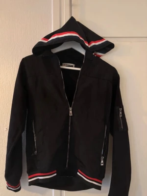 Svart hoodie från Moncler med ränder - Snygg svart hoodie från Moncler med dragkedja framtill, två fickor med dragkedja och en extra ficka på ärmen. Huva med röda, vita och svarta ränder samt matchande detaljer vid ärmslut och midja. Moncler-logga på vänster ärm. Perfekt för en sportig och trendig look.