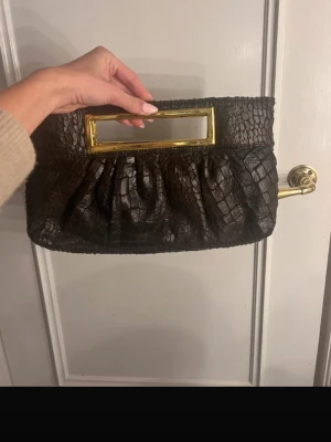 Svart kuvertväska med guldhandtag - Stilren svart kuvertväska i fejkläder med krokodilstruktur och rektangulärt handtag i guldmetall. Väskan har en mjuk, veckad form och passar perfekt när du vill addera en lyxig touch till din outfit.