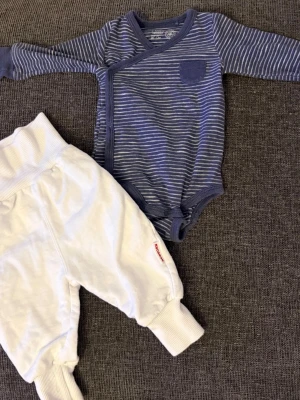 Randig body och vita byxor set - Säljer ett gulligt set från Lindex med en blå och vit randig body med omlottknäppning och liten bröstficka samt ett par mjuka vita byxor med breda muddar. Perfekt för mysiga dagar hemma. Fint skick 