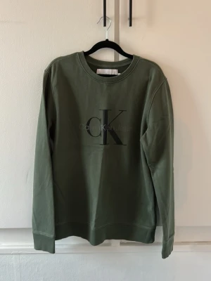 Olivgrön sweatshirt från Calvin Klein - Säljer en olivgrön sweatshirt från Calvin Klein Jeans med stor CK-logga på bröstet. Tröjan har rund halsringning, långa ärmar och ribbade muddar vid ärmslut och nederkant. Perfekt för dig som gillar stilrena och bekväma plagg.