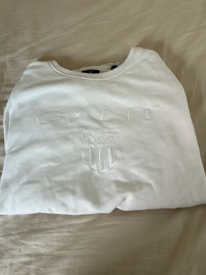 Vit sweatshirt från GANT - Säljer en stilren vit sweatshirt från GANT med broderad logga och sköld på bröstet. Tröjan har rund halsringning, långa ärmar och ribbade muddar vid ärmslut och nederkant. Perfekt för en clean och klassisk look.
