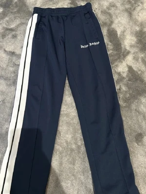 Mörkblå Palm Angels mjukisbyxor - Säljer ett par mörkblå mjukisbyxor från Palm Angels med vita sidoränder och logga på vänster lår. Byxorna har resår i midjan och raka ben. Perfekta för en avslappnad och trendig look.