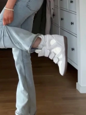 Isabel marant liknande  - Säljer ett par vita sneakers med högt skaft och tre breda kardborreband. Skorna har detaljer i mocka och slät textil, samt en tjock sula i brunt gummi. Perfekta för dig som gillar en sportig och trendig look.