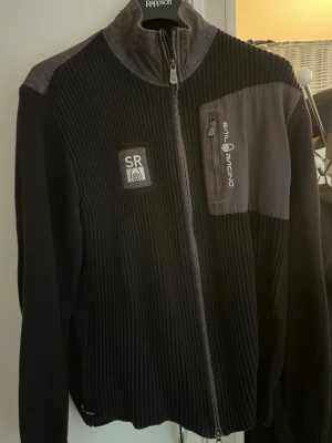 Svart fleecejacka från Sail Racing - Säljer en svart fleecejacka från Sail Racing med ribbad struktur, hel dragkedja och grå detaljer på axlar och bröst. Jackan har en bröstficka med dragkedja och Sail Racing-logga samt SR-märke på vänster sida. Perfekt för dig som gillar sportig stil.
