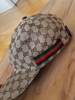 Gucci beige keps med GG-mönster - Säljer en beige keps från Gucci med det klassiska GG-monogrammet över hela kepsen. På sidan finns en grön och röd rand som ger en ikonisk touch. Kepsen har böjd skärm och justerbar rem bak. Materialet är canvasliknande och känns lyxigt.