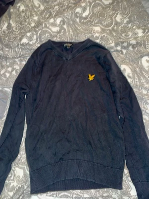 Svart tröja med V-ringning Lyle & Scott - Klassisk svart tröja från Lyle & Scott med V-ringning och den ikoniska gula örnloggan broderad på bröstet. Tröjan har långa ärmar och ribbade muddar vid ärmslut och nederkant. Perfekt till jeans eller chinos för en clean look.