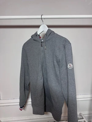 Grå zip hoodie från Moncler - Snygg grå hoodie från Moncler med dragkedja framtill och klassisk huva. Tröjan har ribbade muddar och Moncler-logga på ärmen. Tillverkad i mjukt bomullsmaterial och har diskreta detaljer i rött och blått vid ärmslutet. Hör av dig vid frågor eller funderingar eller vill ha fler bilder🙌