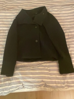 Svart kort kappa med stora knappar - Stilren svart kappa i kort modell med bred krage och två stora knappar framtill. Klassisk och enkel design som passar till många olika outfits. Materialet ser ut att vara tjockt och mjukt, perfekt för kyligare dagar.