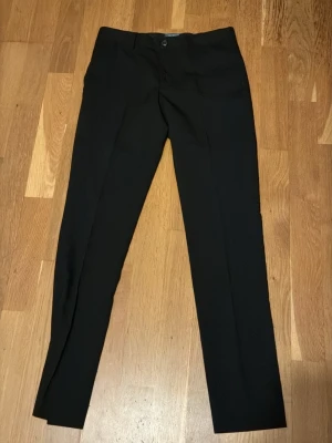 Svarta kostymbyxor från Jack & Jones - Klassiska svarta kostymbyxor från Jack & Jones med rak passform och diskret knappstängning framtill. Perfekta för en stilren look och tillverkade i ett lätt material som ger en snygg siluett. Passar dig som vill ha en enkel och tidlös byxa. Nypris ca 999kr