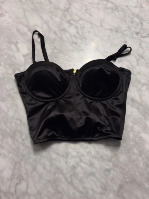 Svart topp  med gulddragkedja - Snygg svart topp med vadderade kupor och justerbara axelband.  har en guldfärgad dragkedja bak och är tillverkad i ett glansigt, satinliknande material. Perfekt för dig som vill ha en trendig och stilren look.