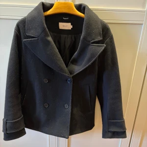 Svart kort kappa från Nelly - Stilren svart kappa från Nelly med dubbelknäppning och bred krage. Kortare modell med två fickor framtill och uppvikta ärmslut. Den är väl använd därav är den nopprig om man kollar närmare. 