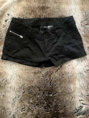 Svarta shorts med dragkedjedetalj - Snygga svarta shorts med låg midja och en cool dragkedjedetalj på ena fickan. Klassisk femficksmodell i bomull med knapp och gylf framtill. Perfekta för varma dagar och enkel att matcha med olika stilar. ( tyvärr har jag tappa knappen )