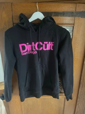 Svart Dirt Cult hoodie med rosa tryck - Svart hoodie från Dirt Cult med stor rosa logga och texten 'San Diego' på bröstet. Klassisk känguruficka framtill och justerbar huva med snören. Perfekt för dig som gillar streetwear och vill sticka ut med färgstarkt tryck.