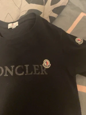 Svart Moncler t-shirt med logga - Snygg svart t-shirt från Moncler med stor logga på bröstet och en mindre patch på ärmen. Klassisk rund hals och korta ärmar. Tillverkad i mjuk bomull som känns skön mot huden. Perfekt för dig som gillar stilrena och exklusiva plagg. Pris kan diskuteras