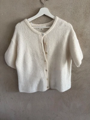 Krämvit kortärmad kofta med knappar - Mysig krämvit kofta med korta ärmar och ribbad struktur. Koftan har rund halsringning och stängs med pärlemorskimrande knappar framtill. Perfekt att slänga över en topp för en chill look. Materialet ser ut att vara mjukt och fluffigt, troligen ull- eller mohairblandning.