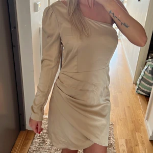 CHAMPAGNE KLÄNNING 36 - Säljer denna fina champagne färgade one shoulder klänningen i satin. Perfekt till fest. Storlek 36. Oanvänd med prislapp kvar på. Säljer för 100 kr. Köparen står för frakten!