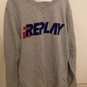 En Replay tröja som är grå med röd blå text på! Har även ett litet R längst med tröjarmen.