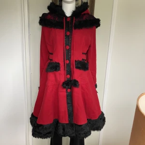 Kappa med fuskpäls  - Aldrig använd, liten i storleken, motsvarar 36/38. Riktiga fickor framtill, midjebandet är ej avtagbart. Detaljer i fuskpäls. Kappan stängs genom att trä knapparna genom en öppning.   Mått i cm: l 97, b 42, ärmlängd 66.