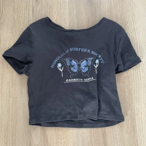 Grå crop top med fjäril - En crop top som är grå med en blå fjäril på 🦋 .tänkte att det var dags att sälja den då jag aldrig använt den.