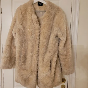 Beige vinterjacka i fuskpäls  - Beige vinterjacka i fuskpäls. Väldigt mysig och varm. Kan tvättas i tvättmaskin. Knäpps med hyskor. Användes förra vintern. Storleken är S, men stor i storleken.  Materialet är polyester. 