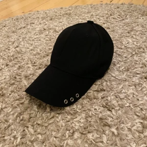 Svart keps  - Svart keps med silver hål på vänstra sidan av skärmen🖤regleringsbart band på baksidan.  Aldrig använd!