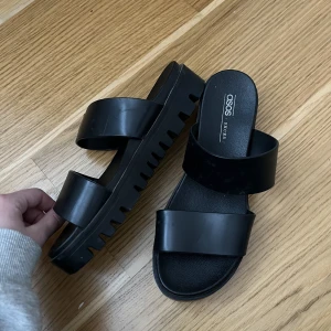 Svarta tofflor - Svarta tofflor/sandaler från ASOS. De är i gummi/plast. Använda några få gånger då min vrist är för hög så de sitter inte bra på min fot. Storlek 39