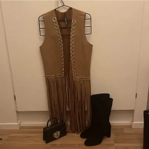 Boho vintage-chic mockaväst med fransar - En väst i mockaimitation med fransar nedtill och guld-detaljer. Vintage och Boho Western Vibes. Helt ny i perfekt skick, aldrig använd. Köpt för Cochella musikfestival. Stl onesize M-L, köpt på asos från märket Lily white. Nypris 599! Samfraktar! 