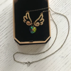 Halsband med glittrande hjärta & vingar - Halsband i metall och plast från Sugar Bunny Shop. Fått i present men det är inte min stil alls, sparsamt använd men det finns visst slitage/tecken på användning på materialet. Asken ingår. Köpare betalar frakt.