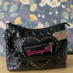 Betsey Johnson Handväska - Handväska från Betsey Johnson. Quiltade hjärtan och hjärtdetaljer.  Guldig tag som går att ta av. Axelremmen är en svart plastkedja.  I jättefint skick. Mått: 42 cm lång och 30 cm hög