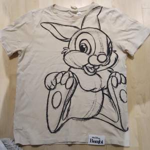 En enkel ljus beige T-shirt med ett skissat tryck av Stampe från Disney filmen Bambi
