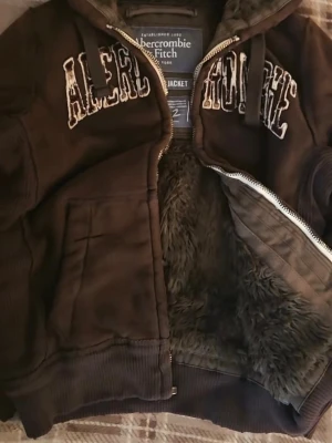 Brun hoodie från Abercrombie & Fitch - Tar bud; Mysig brun hoodie från Abercrombie & Fitch med dragkedja och stor huva. Insidan är fodrad med mjuk fuskpäls för extra värme. Stora broderade bokstäver på bröstet och två fickor framtill. Perfekt för kalla dagar. 