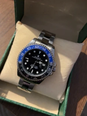 Klocka - Snygg klocka med blå och svart vridring, svart urtavla och silverfärgad länk i rostfritt stål. Klockan har tydliga index, datumfönster och klassisk design. Perfekt för dig som gillar exklusiva accessoarer.