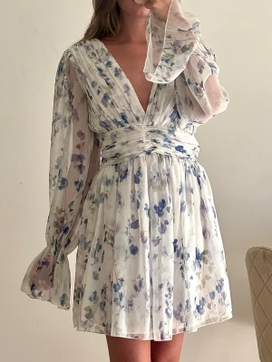 Blommig kortklänning med volangärm - Söt sommarklänning i vitt med blått och grönt blommönster. Klänningen har v-ringning, markerad midja och långa, vida ärmar med volangdetalj vid ärmslut. Tillverkad i ett lätt och luftigt material som ger en romantisk känsla.