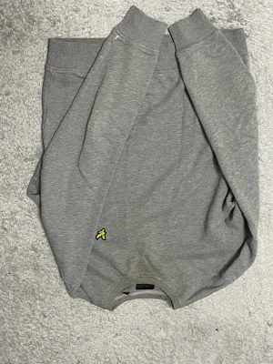 Grå sweatshirt från Lyle & Scott - Snygg grå sweatshirt från Lyle & Scott med klassisk rund halsringning och den ikoniska gula örnloggan broderad framtill. Tillverkad i mjukt bomullsmaterial och har ribbade muddar vid ärmslut och nederkant. Perfekt för en avslappnad och stilren look.