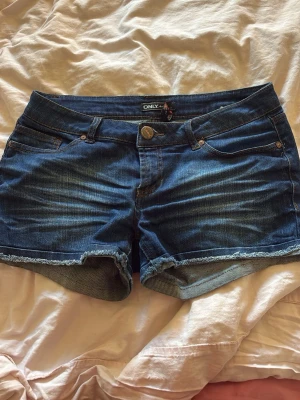 Blå jeansshorts från ONLY - Säljer ett par klassiska blå jeansshorts från ONLY med råa kanter och femficksmodell. Shortsen har normal passform och är tillverkade i slitstark bomull med snygga slitningar och detaljerade sömmar. Perfekta för varma dagar och enkel att matcha. De är lågmidjade 