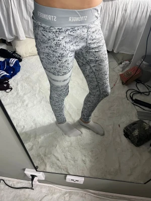 Gråmönstrade leggings från Stronger - Säljer ett par snygga leggings från Stronger i grått och vitt med abstrakt mönster och två vita ränder på ena låret. Med resår där det står 'STRONGER'. Perfekta för träning eller chill dagar.