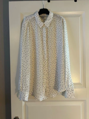 Vit prickig blus med krage - Stilren vit blus med svarta prickar och klassisk krage. Blusen har knäppning framtill och långa ärmar med manschetter. Materialet ser lätt och luftigt ut, perfekt för en trendig och avslappnad look.