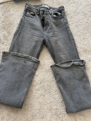 Gråa bootcut jeans - Säljer ett par gråa jeans med rak passform och uppvikta benslut. Jeansen har klassisk femficksdesign och bälteshällor. Materialet är denim i bomull och de har en snygg tvättad look som passar till det mesta.