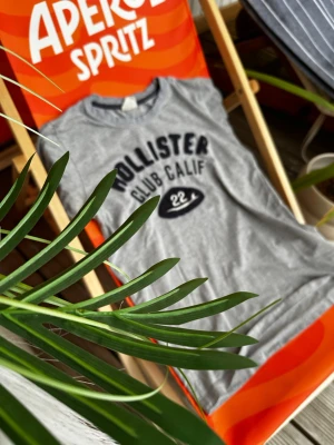 Grå Hollister t-shirt med tryck - Snygg grå t-shirt från Hollister med svart text 'Hollister Club Calif' och en liten detalj med siffran 22 framtill. Klassisk rund halsringning och korta ärmar. Perfekt till jeans eller shorts för en avslappnad look.
