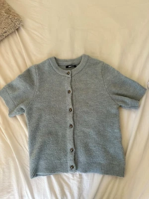 Ljusblå kortärmad kofta Gina Tricot - Supersöt ljusblå kofta från Gina Tricot med korta ärmar och rund hals. Koftan har ribbade muddar och knäpps med silvriga knappar framtill. Perfekt att slänga över en topp för en chill look.