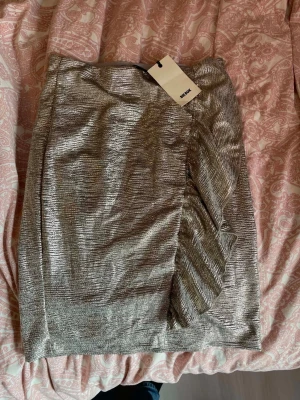 Helt ny med prislapp, S, Glittrig silverkjol från BikBok - Säljer en glittrig silverfärgad kjol från BikBok i kort modell. Kjolen har en snygg omlottdetalj framtill och är perfekt för dig som vill sticka ut. Materialet är skimrande och ger en festlig känsla. Supersnygg att styla med en enkel topp eller crop top.
