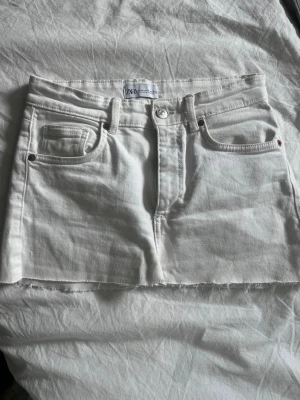 Vit jeanskjol från Zara - Säljer en vit kort jeanskjol från Zara med rå kant nertill och klassiska fem fickor. Kjolen har knapp- och dragkedjestängning framtill och är gjord i ett stadigt bomullstyg. Perfekt för dig som gillar en enkel men snygg stil.