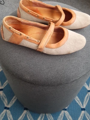 Beige Timberland  ballerinaskor med läderdetaljer - Snygga ballerinaskor från Timberland i beige textil med ljusbruna läderdetaljer och rem över foten. Skorna har rund tå och dekorativa öljetter på sidorna. Perfekta för en avslappnad och trendig stil.