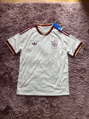 Vit Spanien Adidas fotbollströja - Säljer en vit fotbollströja från Adidas med Spaniens landslagsemblem på bröstet. Tröjan har vinröda och gula detaljer vid krage, ärmslut och axlar samt Adidas klassiska ränder. Tillverkad i lätt och skönt material, perfekt för träning eller match.
